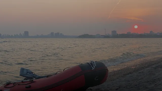 A red and black inflatable boat with a small motor rests on a dark pebbled shore, with a hazy city skyline and low red sun across the water.