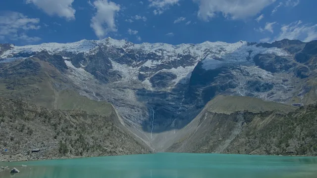A turquoise lake lies below steep, rocky mountains capped with snow and ice, under a blue sky with a few clouds.