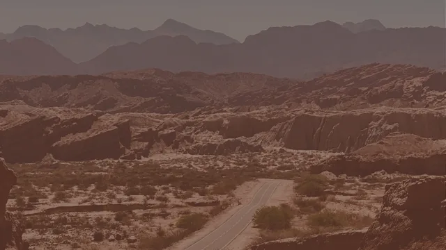 A paved road with lane markings winds through dry scrubland between eroded red-brown rock formations, with hazy mountains beyond.