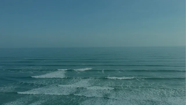 Looking down on rows of small waves with white foam on blue-green water, under a pale blue-grey sky.