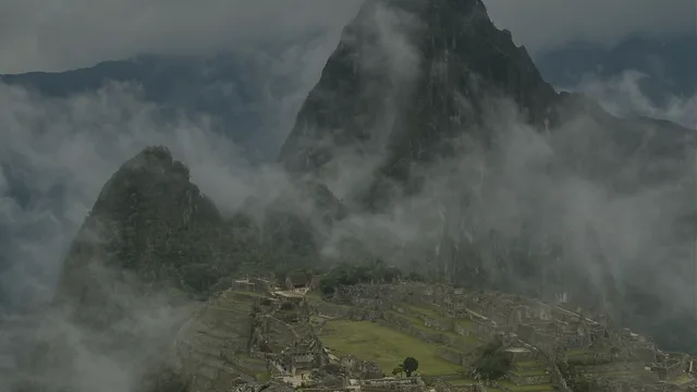 Stone ruins and green terraces sit below steep, forested peaks, with low cloud drifting through the mountains.