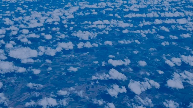 Aerial view of small white clouds scattered above a patchwork of fields, all in deep blue tones.