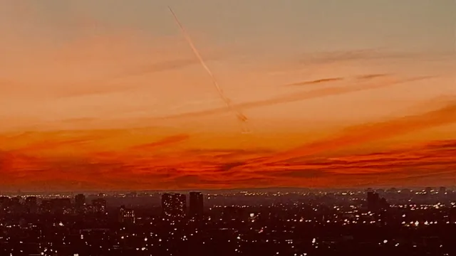A thin white contrail crosses an orange and red sky above a dark city with scattered lights.