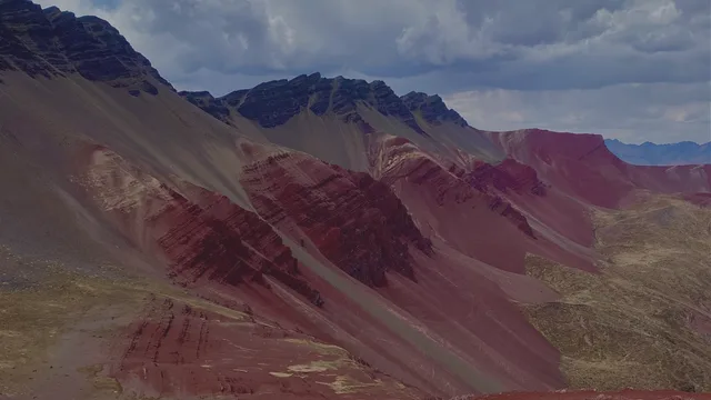 Steep mountain slopes banded in red, maroon and grey rise under a cloudy blue sky, with a dark rocky ridge along the top.